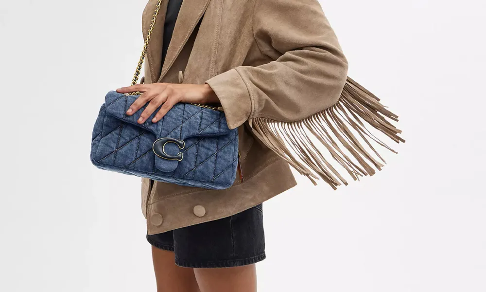 A cropped model in a tan fringed jacket and black shorts holds a quilted blue handbag with a gold clasp, showcasing a chic fashion look.