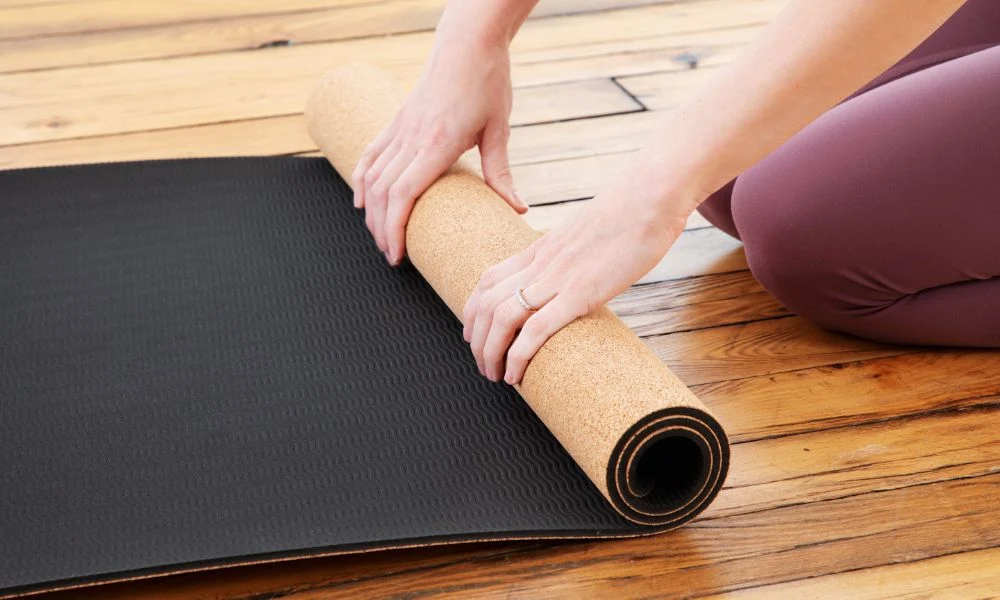 A person in plum leggings rolls a black yoga mat across a wooden floor, revealing its tan underside in a calm moment of workout preparation.