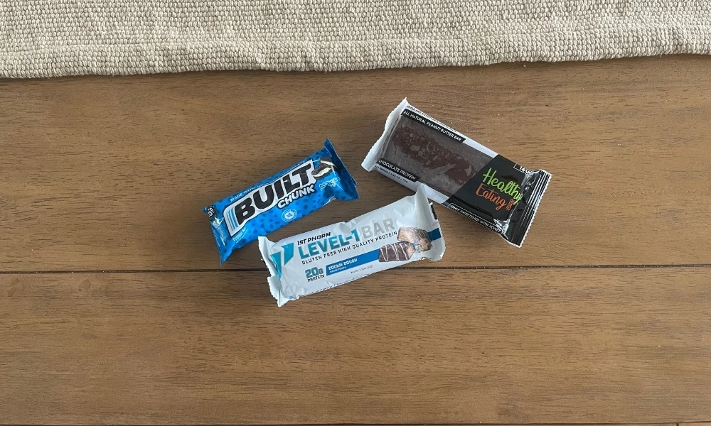 Three packaged protein bars—two in blue wrappers and one dark brown—lie on a wooden tabletop below a strip of textured fabric, suggesting a simple snack selection.