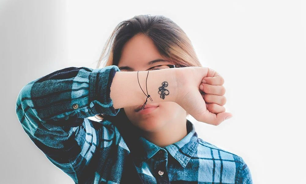 A young woman in a blue plaid shirt hides part of her face behind her arm, showing a small wrist tattoo and bracelet in a playful studio portrait.