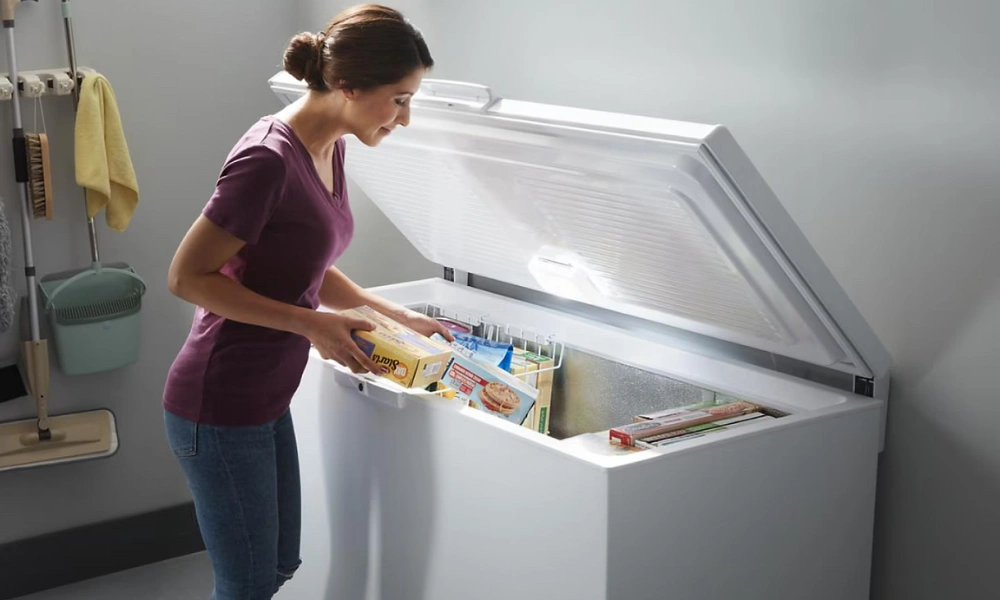A woman in a purple shirt opens a white chest freezer and reaches for a boxed frozen meal, in a clean, well-lit utility room.
