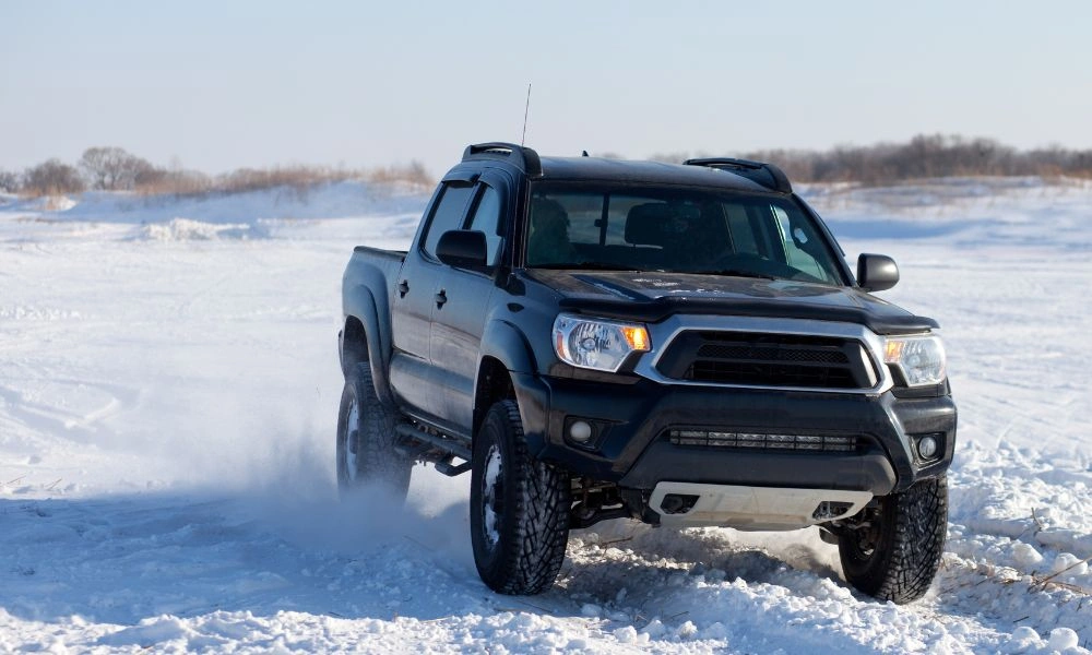 A dark blue off-road pickup truck drives through deep snow, kicking up powder beneath a clear winter sky; the scene feels rugged and adventurous.