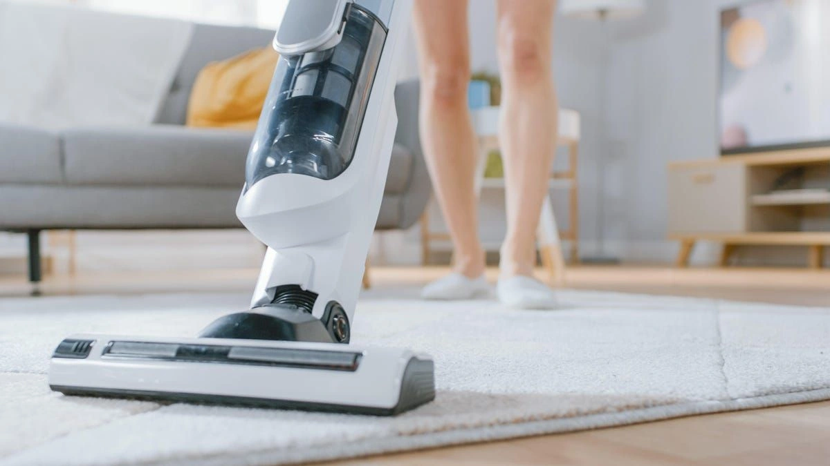 A person vacuums a light rug with a white cordless upright vacuum in a bright living room, creating a calm, clean home atmosphere.