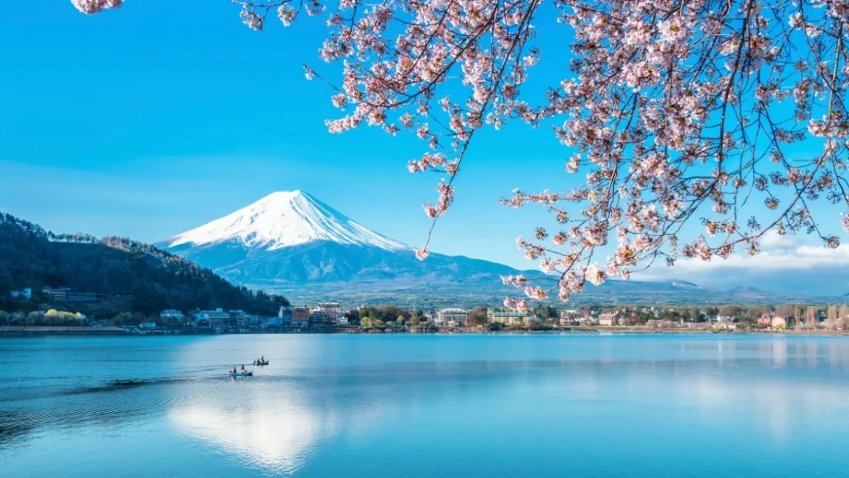 Pink cherry blossoms frame snow-capped Mount Fuji across a calm blue lake, with a small boat and lakeside town beneath a clear sky; serene spring scene.