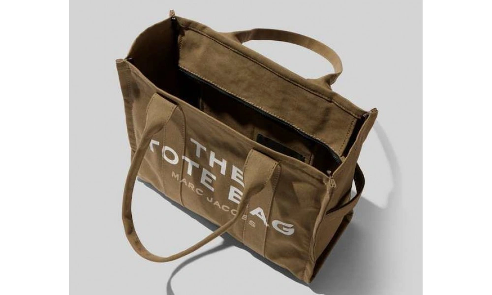 Olive-brown canvas tote bag sits open, showing its dark interior; long handles and white “THE TOTE BAG” lettering give it a casual, utilitarian look.