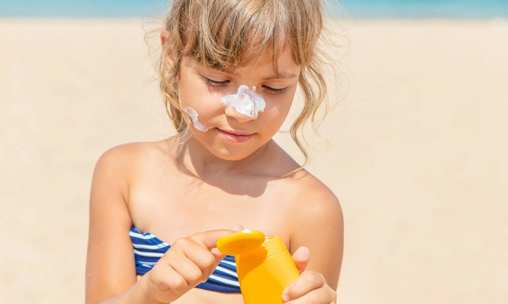 A young girl in a blue striped swimsuit applies sunscreen to her nose at the beach, holding a yellow bottle in bright sunlight. Her expression is calm and focused.