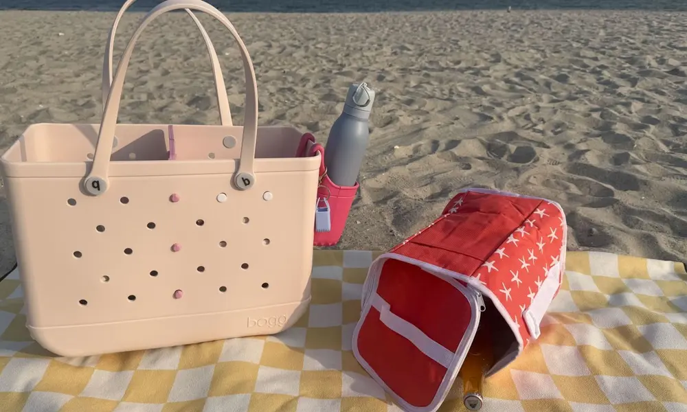 A blush-pink perforated tote, water bottle, and red star-patterned beach bag rest on a yellow-and-white picnic blanket by the sunny, rippled shore.