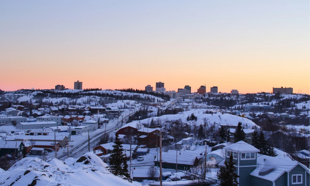 Snow-covered residential neighborhood and distant city skyline at sunset, with pastel pink and orange light on the horizon; a quiet, wintry scene.