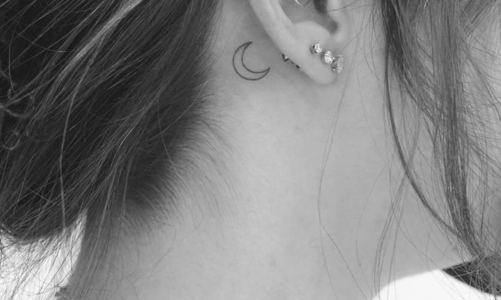 Minimalist crescent moon tattoo behind the ear.