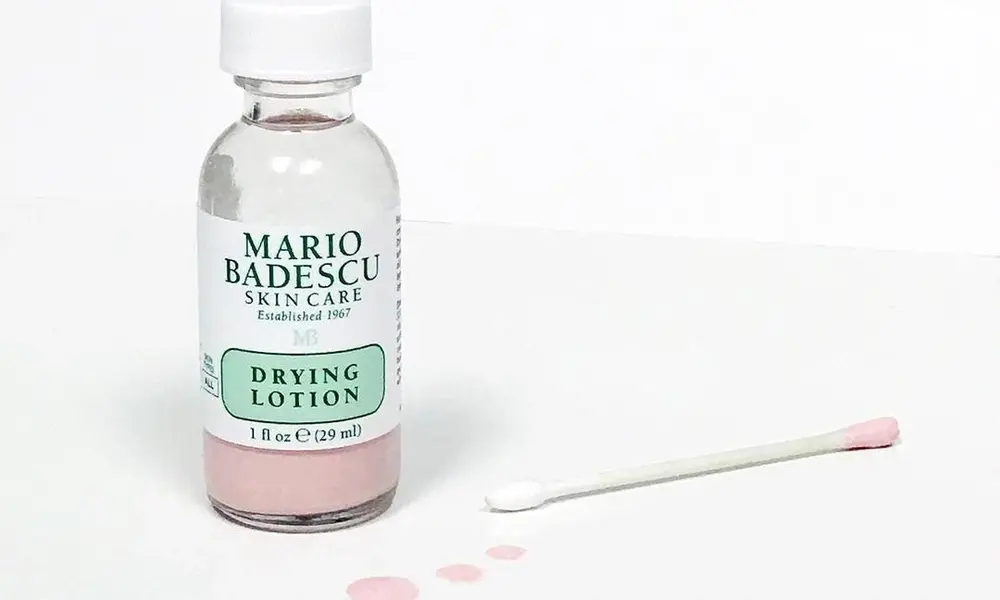 A Mario Badescu Drying Lotion bottle stands beside a cotton swab on a white surface, with pink lotion visible in the bottle and small drops nearby.