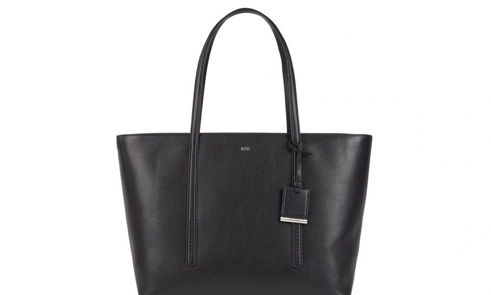 Black leather tote bag with a structured, wide silhouette, long double handles, and a hanging rectangular tag, photographed against a plain white background.