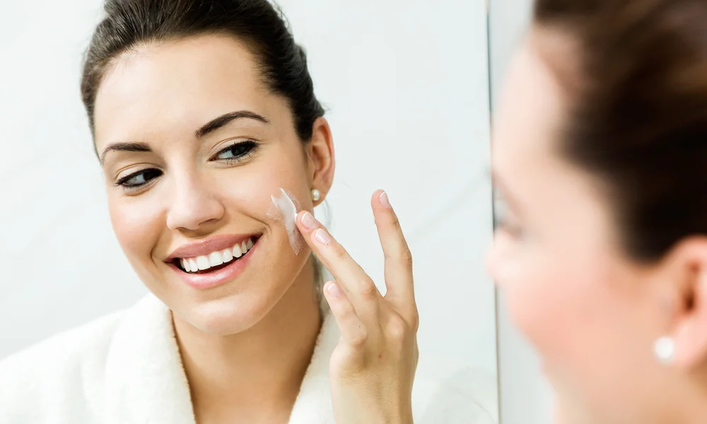 Smiling woman in a white robe applies facial cream to her cheek-best skincare products for dry skin