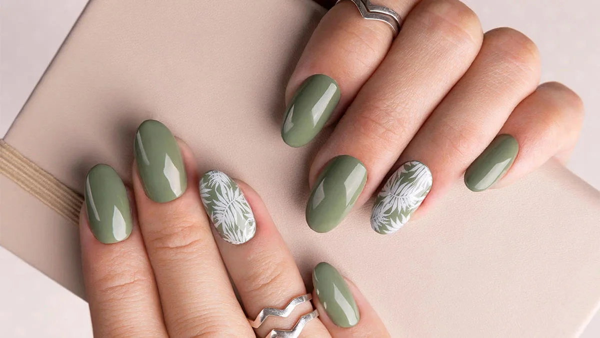 Two hands display a sage-green manicure with glossy nails and white botanical leaf accents, complemented by silver rings against a soft neutral background.
