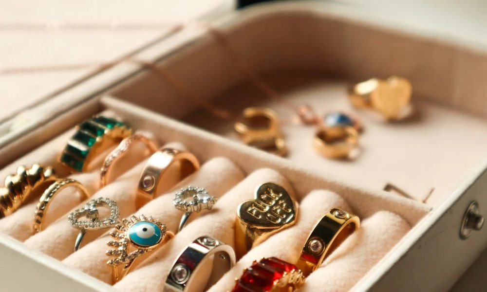 A jewelry box holds gold rings, earrings, and gemstone pieces beside a satin ribbon, creating a warm, elegant display on a weathered blue surface.