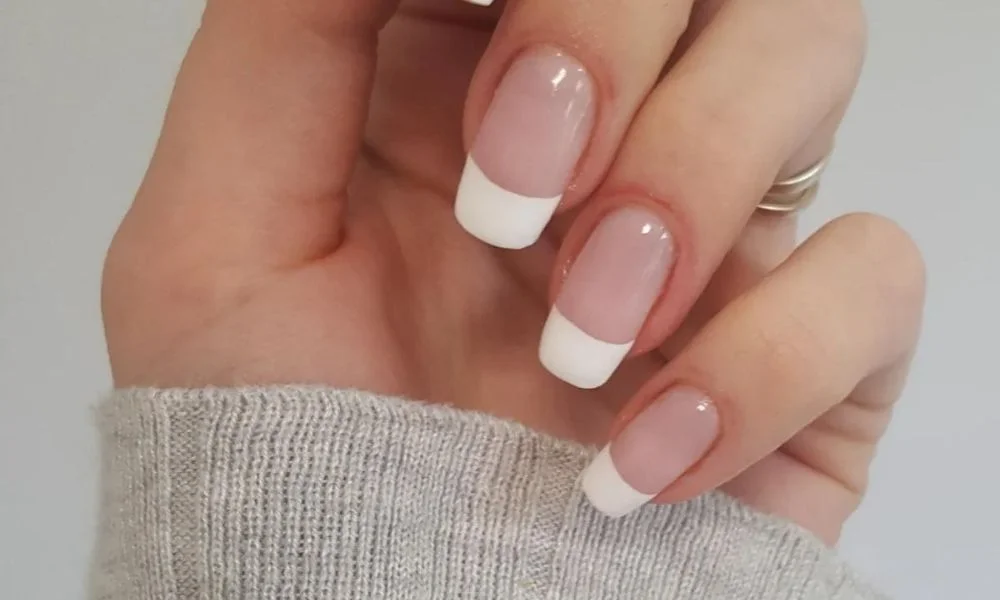 Close-up of a hand with glossy pink French-tip nails and a silver ring, resting above a gray knit sweater against a neutral background; polished, elegant mood.