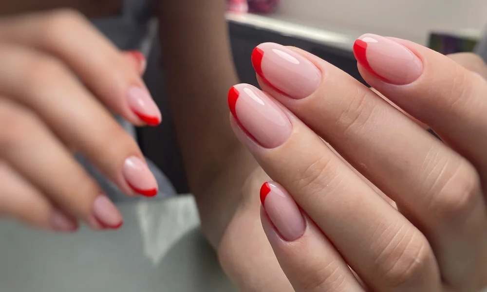 Close-up of hands showing glossy pale pink French-tip nails with bold red edges, displayed after a manicure in a salon; polished, elegant mood.