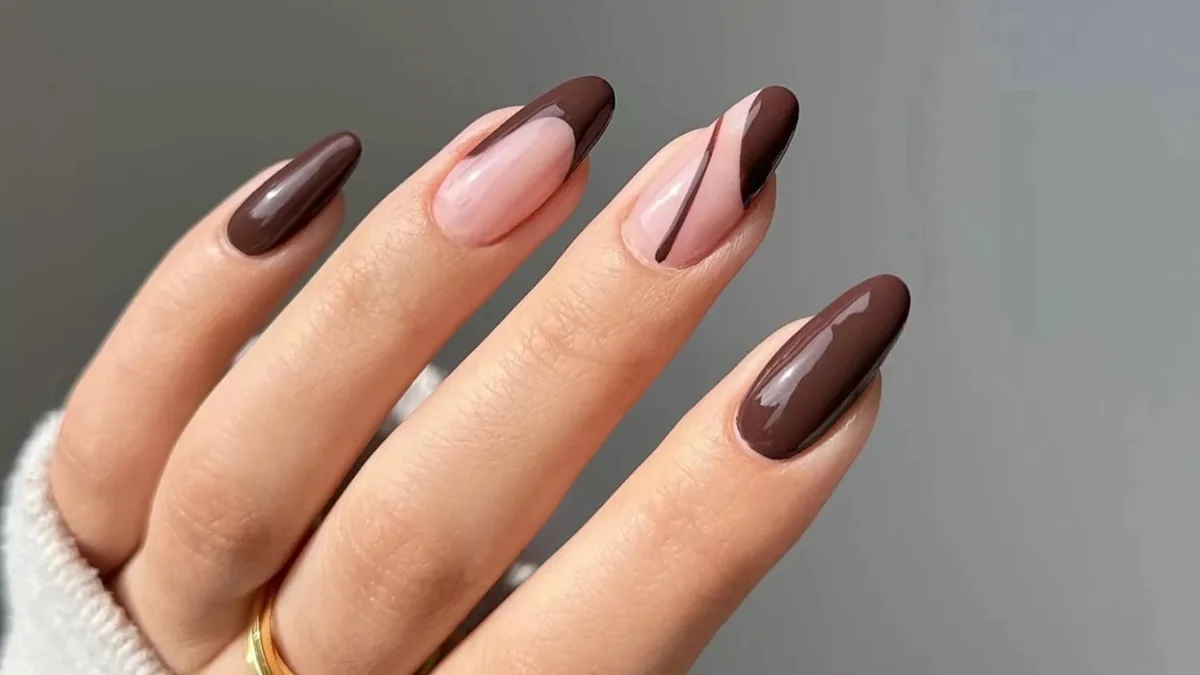 A hand displays long almond-shaped nails with glossy chocolate-brown and blush-pink polish, accented by fine dark lines; elegant manicure against a gray background.