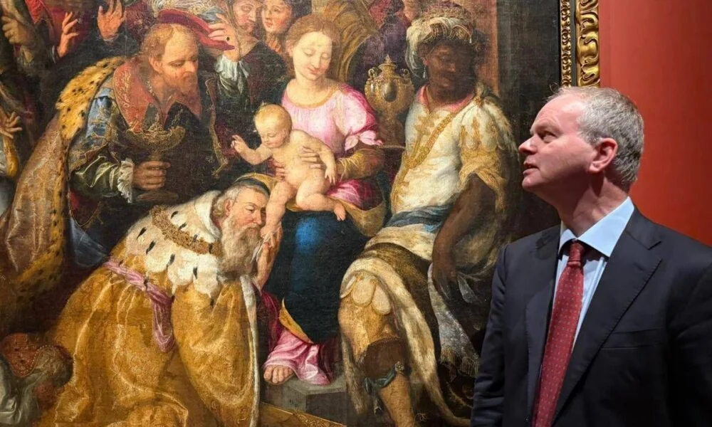 A man in a dark suit studies a crowded Renaissance painting of the Madonna and child, kneeling figures, and richly dressed onlookers; contemplative museum scene.