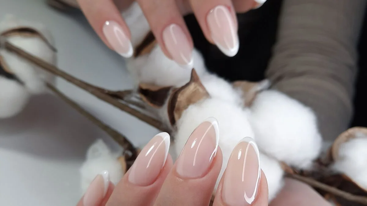 Close-up of manicured hands with glossy pink nails gently touching fluffy white cotton bolls on a branch, creating a soft, delicate beauty mood.