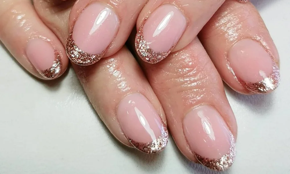 Close-up of manicured hands with glossy blush-pink almond nails and sparkling rose-gold glitter French tips, giving an elegant, festive look.-French Manicure Ideas