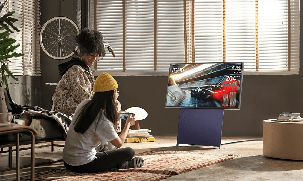 A smiling woman in a yellow beanie and a man sit on a rug watching a large TV in a sunlit living room, enjoying a relaxed moment together-Best Samsung TVs for a Luxury Home