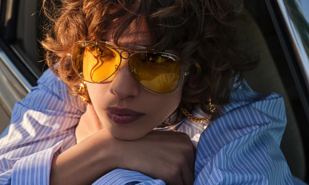 A fashion portrait of a woman reclining in a car, wearing amber-tinted sunglasses and a blue striped shirt, with a relaxed, introspective mood.
