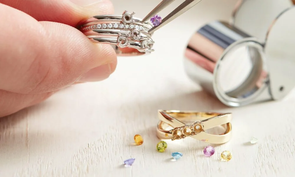 A jeweler’s fingers inspect a silver gemstone ring with tweezers beside a magnifying loupe, gold ring, and scattered gems, suggesting careful repair.