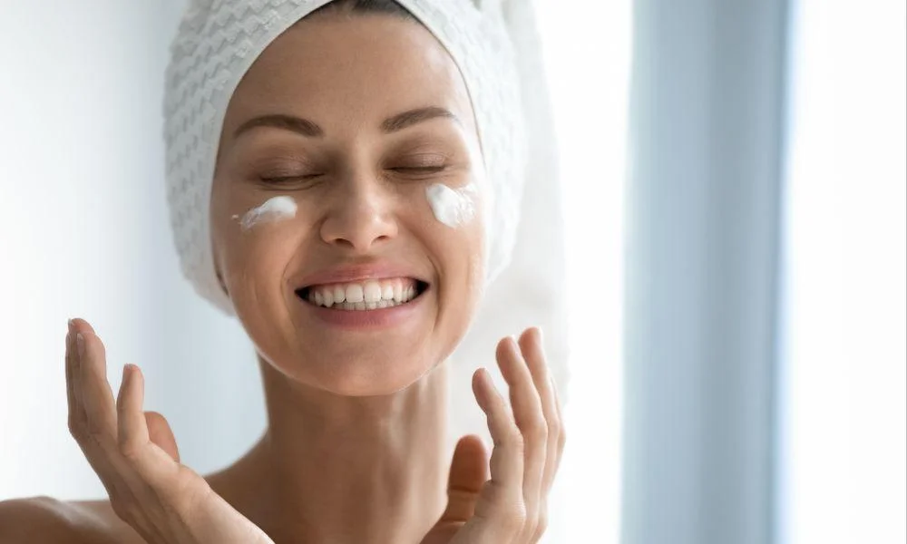 A smiling woman in a white towel turban applies cream beneath her eyes, eyes closed and hands raised, enjoying a calm skincare routine in bright light.