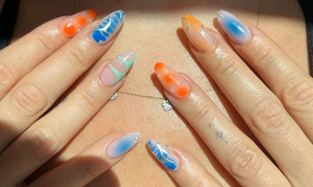 Two hands display long, glossy multicolored nail art with swirls and raised accents over bare skin; warm lighting gives the close-up a playful, summery feel.