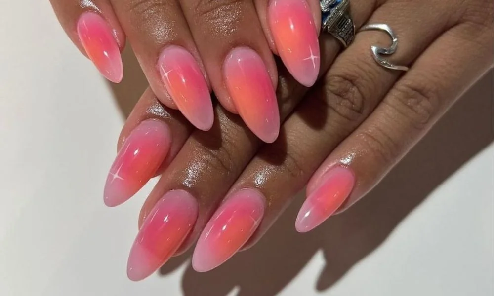 Close-up of hands with long, almond-shaped coral-pink ombré nails and glossy finishes, layered over each other beside a silver ring; polished, glamorous style.