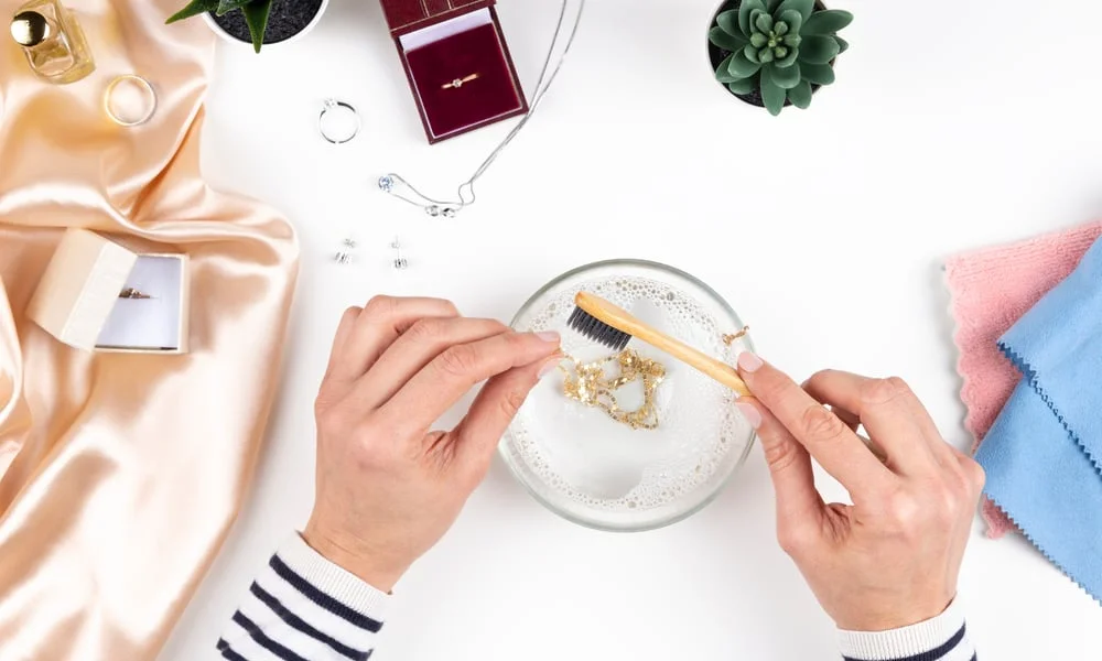 Overhead view of hands gently cleaning gold jewelry in a glass dish with a small brush, surrounded by satin, jewelry boxes, plants, and cloths.