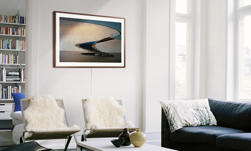 A bright, serene living room features two white chairs, a black sofa, tall windows, and framed art of a curved staircase over water.