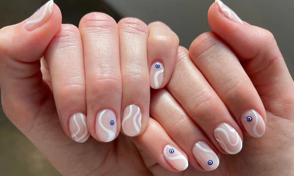 Close-up of two hands with pale pink manicures, decorated with white flowing waves and tiny blue-centered dots; a polished, playful nail-art display.