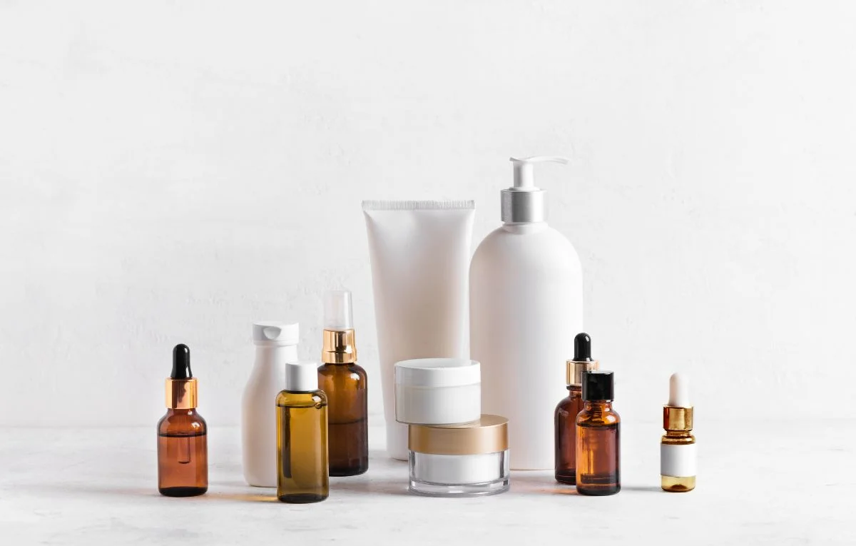 A minimalist skincare collection of amber dropper bottles, clear jars, a white tube and pump dispenser arranged on a pale stone surface, conveying calm simplicity.