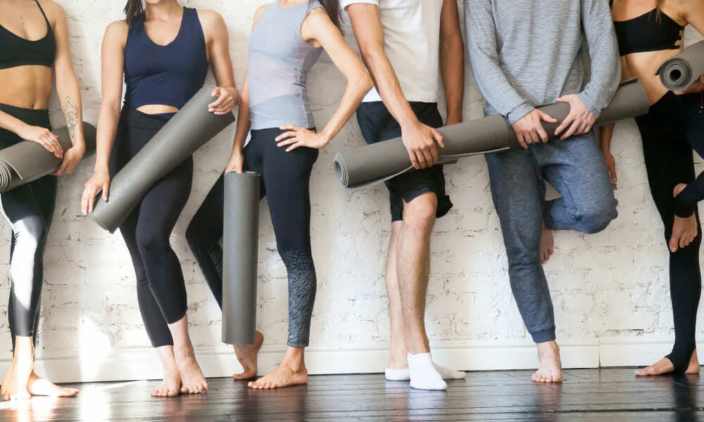 A group of adults in workout clothes hold rolled yoga mats against a white brick wall, ready for a relaxed, social fitness class.
