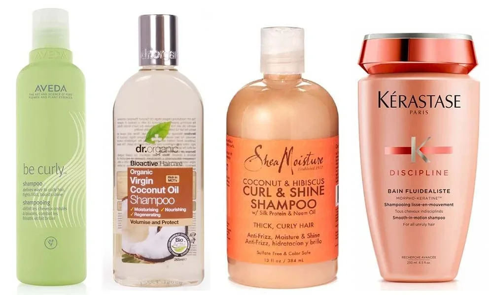 Four hair-care bottles stand side by side: green Aveda Be Curly shampoo, clear Dr. Organic coconut oil shampoo, orange SheaMoisture curl-and-shine shampoo, and pink Kérastase Discipline Bain Fluidealiste. The clean, bright product lineup feels polished and natural.
