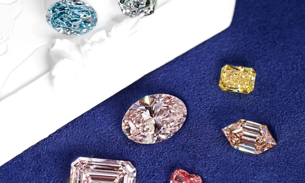 Luxury Diamonds featured in a top-down view of an assortment of fancy-colored loose gemstones, including pink, yellow, blue, and clear diamonds in various cuts, arranged on a deep blue felt surface alongside a carved white pedestal.