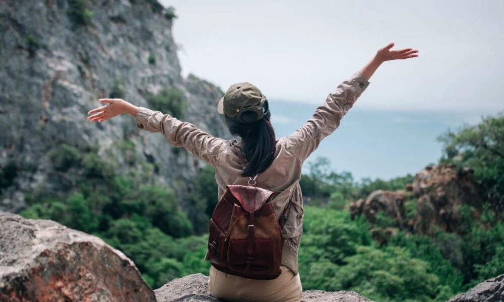 A hiker with a backpack stands on a rocky overlook, arms spread wide toward a green valley and distant horizon, conveying freedom and joy.