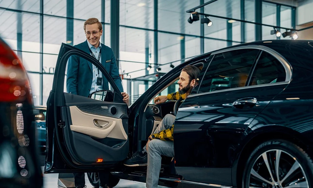Customer sitting inside a open-door black sedan while talking with a sales representative inside a dealership