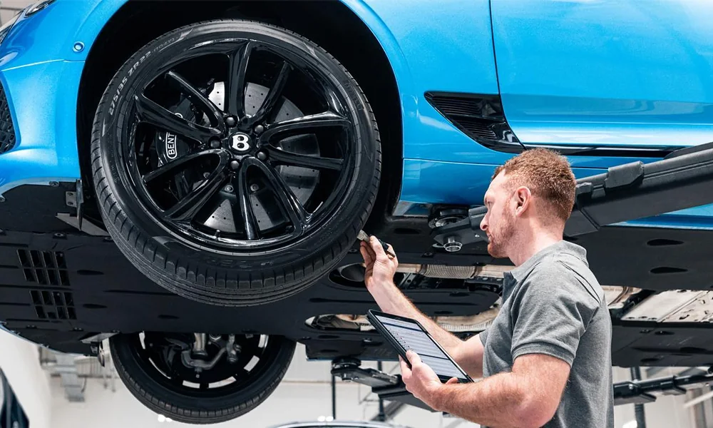 Technician using a digital diagnostic tablet while inspecting the wheel and brake assembly of a blue vehicle on a lift