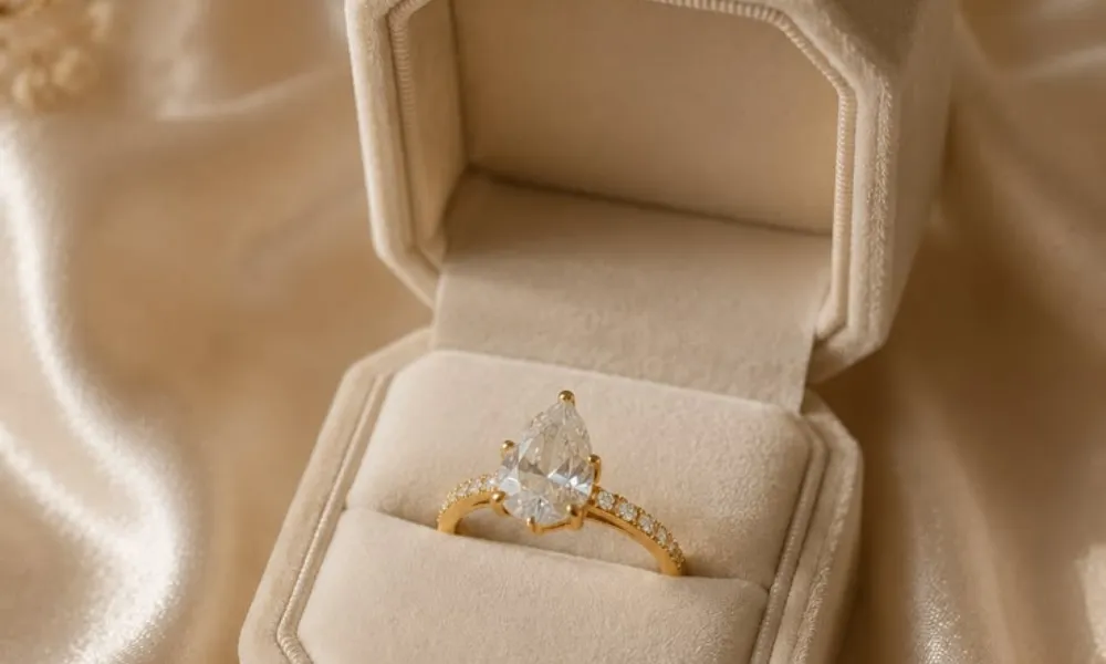Luxury Engagement Ring Box featured in a close-up photo of a pear-cut diamond ring on a pavé gold band resting inside an open beige velvet hexagonal ring box on silk fabric.