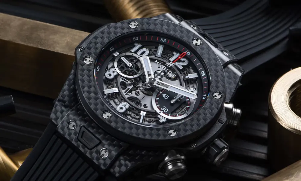 Luxury of Time by Diamonds International featured in a close-up photo of a black carbon fiber Hublot Big Bang skeleton chronograph watch with a black rubber strap resting on brass fittings. 