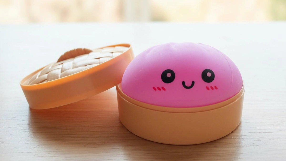Bright pink smiling dumpling-shaped NeeDoh sensory toy sitting inside a plastic bamboo steamer container
