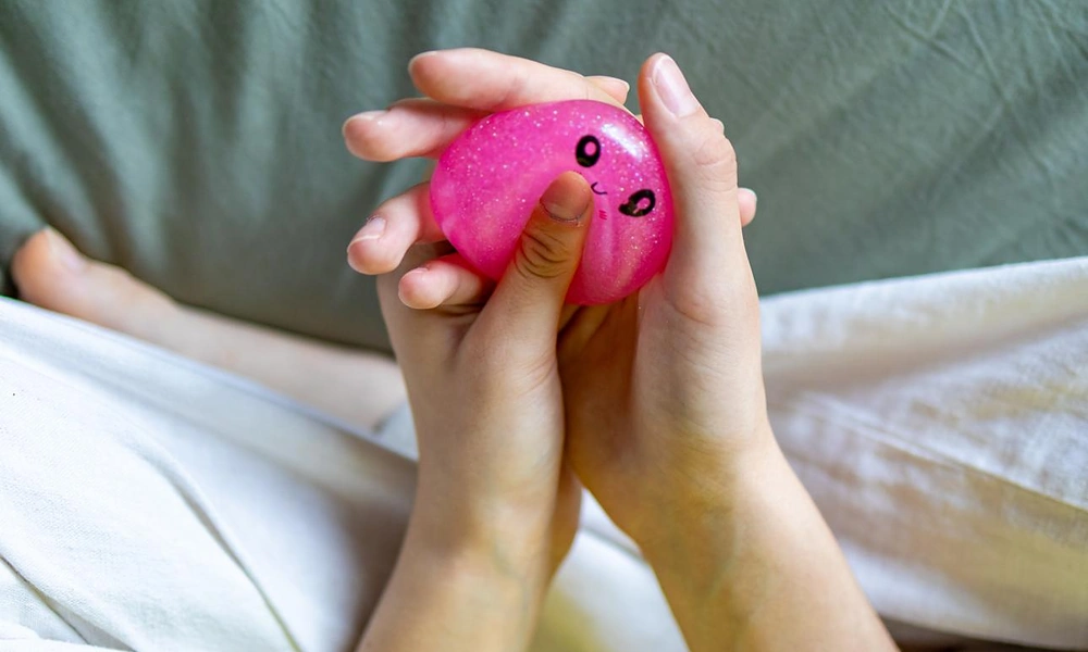 Hands pressing down on a glittery pink smiling stress toy showing pliable material deformation