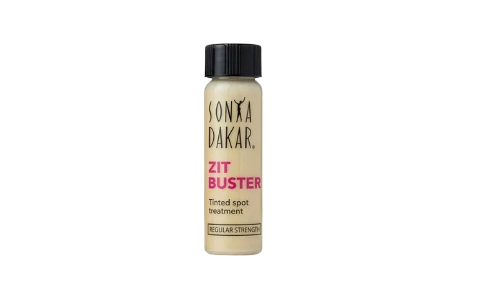 A small beige Sonya Dakar Zit Buster tinted spot treatment bottle with a black cap stands against a solid black background, in a clean, minimalist product image.