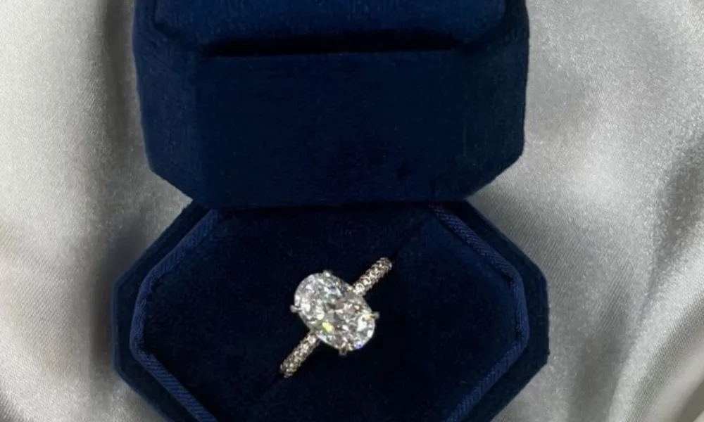 Luxury Engagement Ring Box featured in a top-down photo of an oval-cut solitaire diamond ring on a pavé band inside an open hexagonal navy blue velvet ring box resting on white silk.
